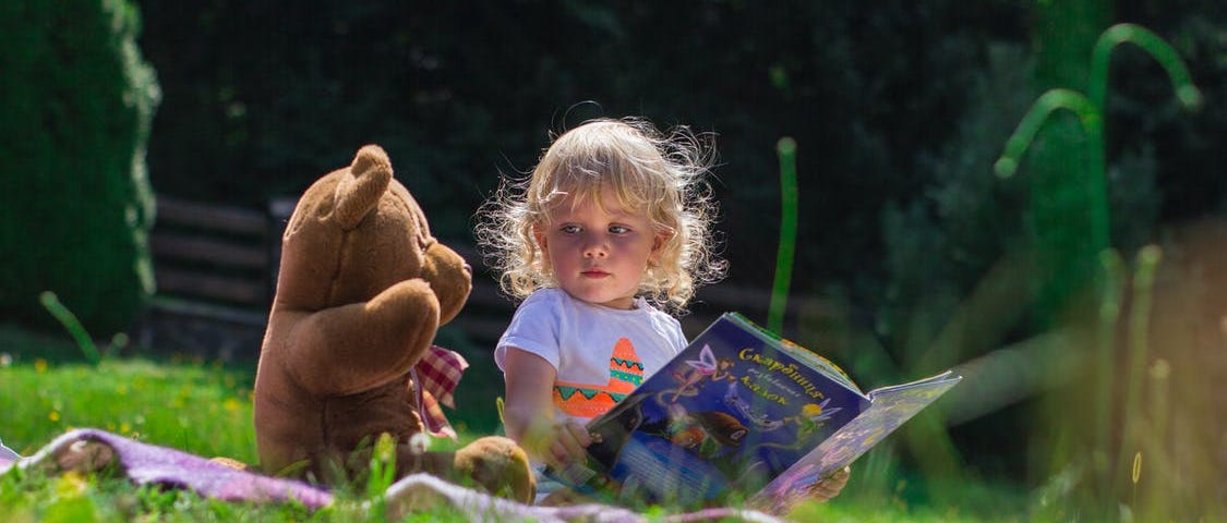 uma foto de uma criança de pele clara e cabelos loiros cacheados e curtos sentada em um pano, que está na grama, ao lado de um urso de pelúcia. a criança segura um livro e olha para o urso de pelúcia.