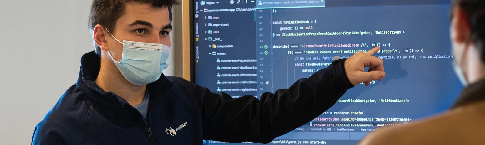 Man with a face mask on showing someone how to code on a big screen.