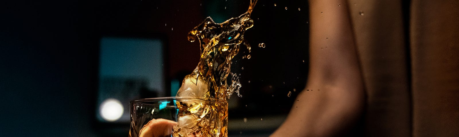 An arm extends down from the upper right to hold a medium-sized glass. Alcohol pours from above, splashing amber-colored fluid into the glass (with some spilling out).