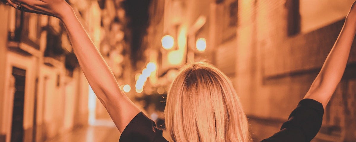 woman with long blonde hair wearing a black shirt standing with arms up in the air looking down street