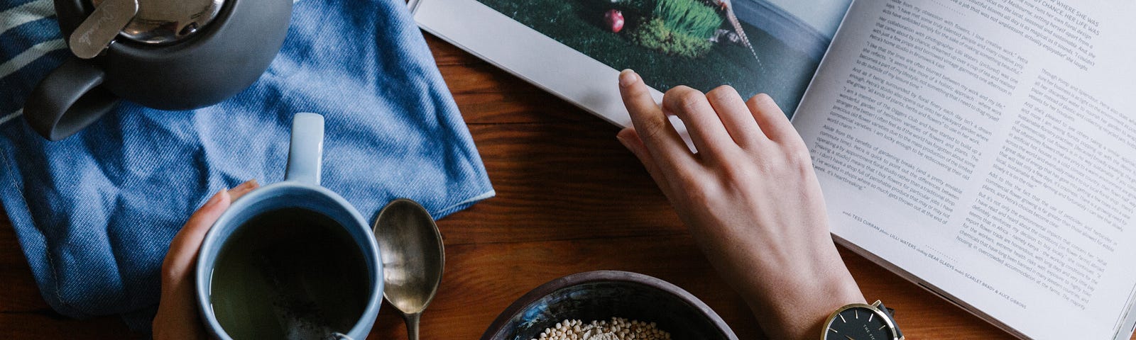 A table with a magazine, coffee pot, bowl of cereal sitting on it. You can also see a persons arms and hands, they are holding a coffee cup and their other hand is resting on the magazine.