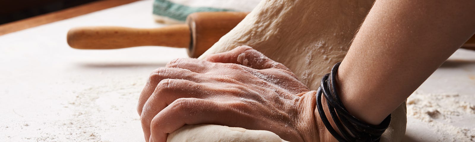 Hands kneed dough on a floured countertop with rolling pin in background.