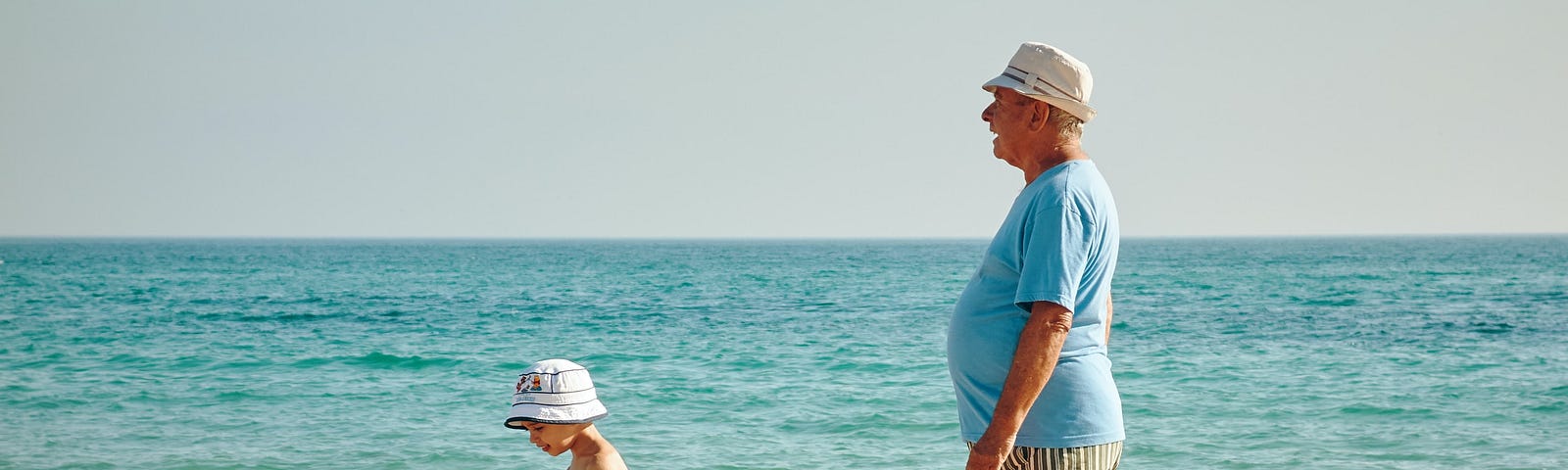 Young child and older man on the beach. Both white with hats. In the background is the ocean.