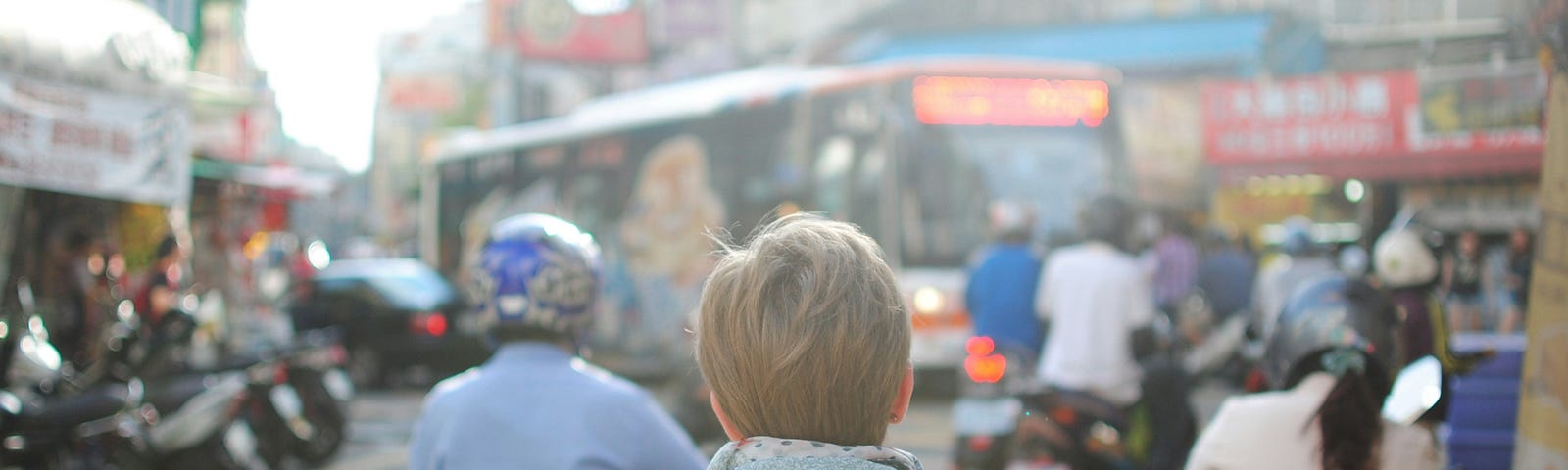 A person stands with their back to the camera, a small rucksack on their back, looking out at a chaotic city road junction full of mopeds and buses