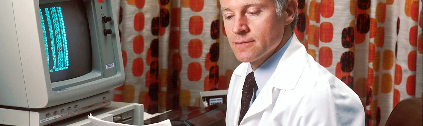 A man in a white medical coat looks at papers, sitting in front of a large, old computer console (circa 1980s). The patterned drape in the background suggests the 1970s or early 1980s.