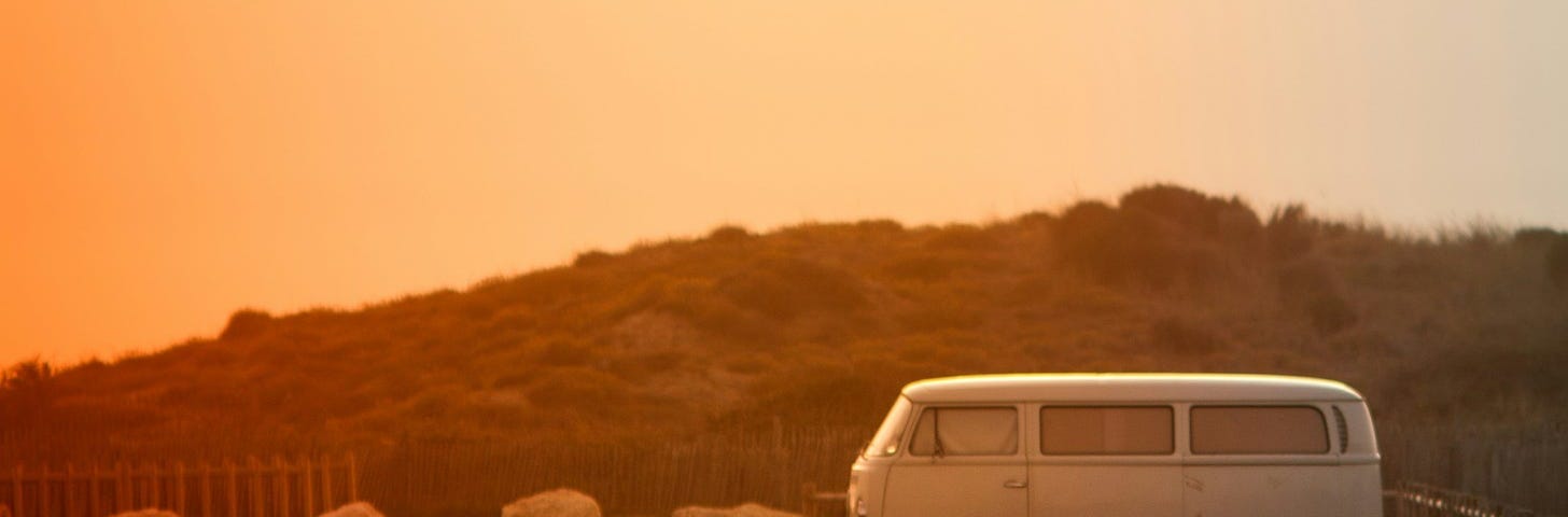 A hazy orange sunset with a rustic VW van parked on an open road.