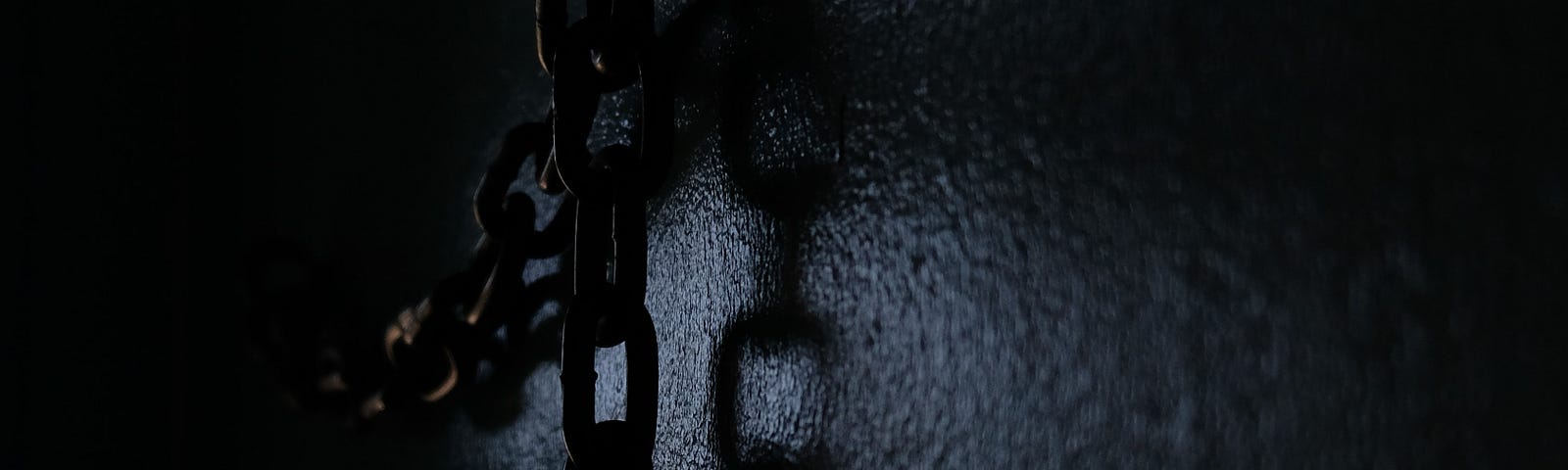 A chain hanging from a hook on a dark wall, casting a shadow.