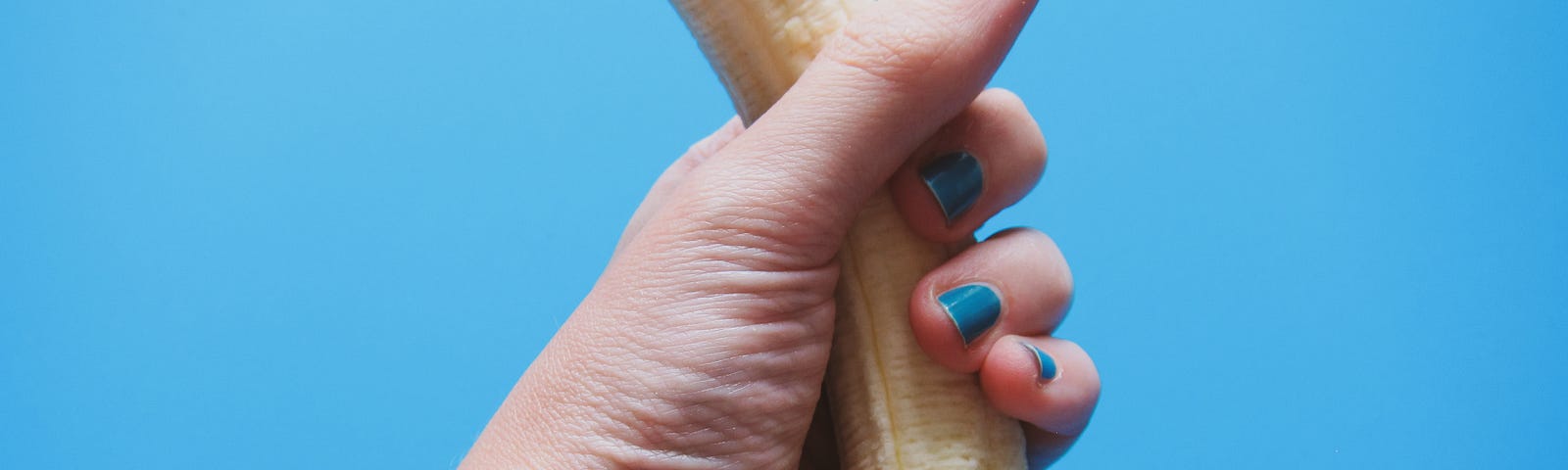 A pink-toned human hand and wrist are visible. There is a hollow star tattoo near the inner wrist. The fingernails are short and painted blue. The hand is clenched around an upright, unpeeled banana. The photo is taken against a blue backdrop.