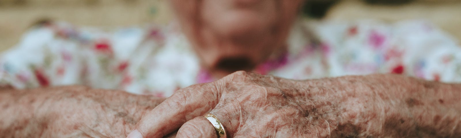 A close up of very aged hands, clasped in front of an elderly woman wearing a white and pink floral blouse.