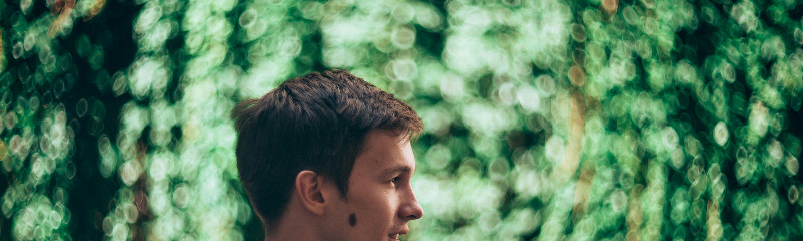 A young man turns his face in profile and looks to his left in front of a swirling green background