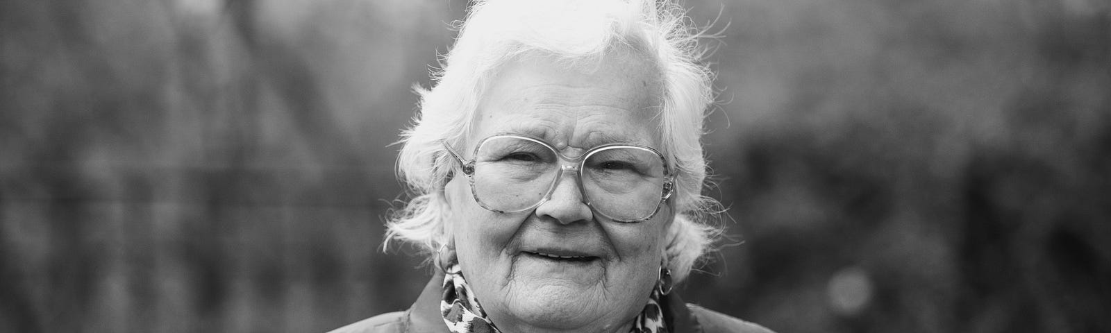A black and white photo of an old lady with gray hair, glasses and a sly grin
