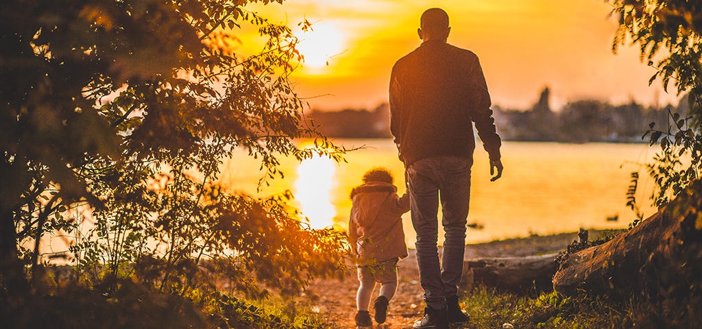 Father and daughter walking near river while sun is setting down