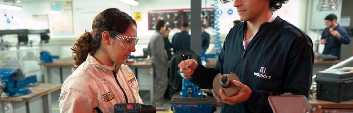 Photo of two students in a workshop wearing safety goggles and jumpsuits. The student on the left is holding a drill. The student on the right is holding materials for the project on the table between them.