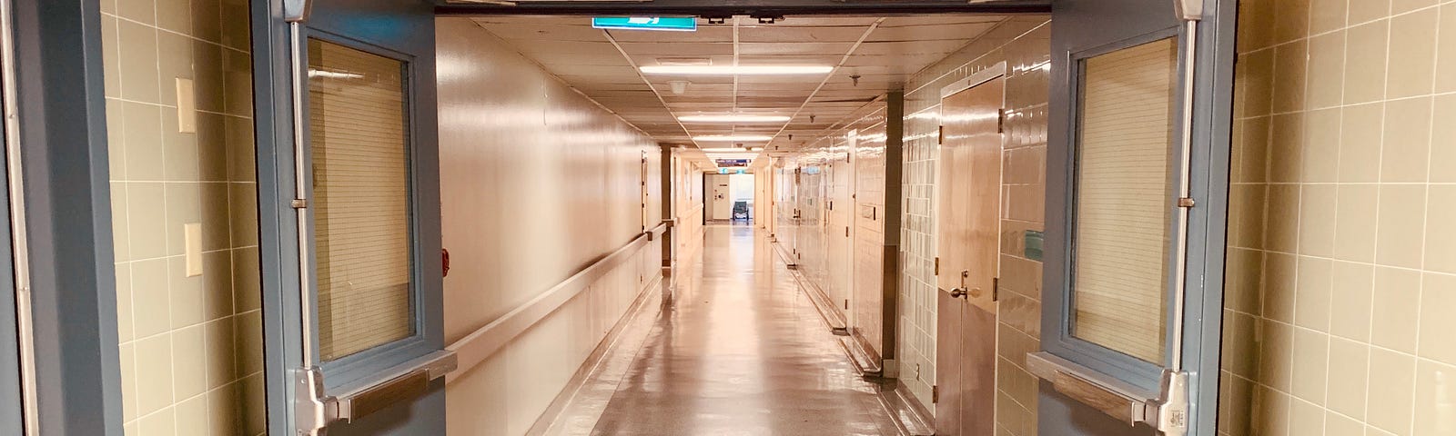 A hospital doorway is opened. Past the doorway is a long hallway with beige walls. There is an emergency exit sign in the top left corner of the hallway.