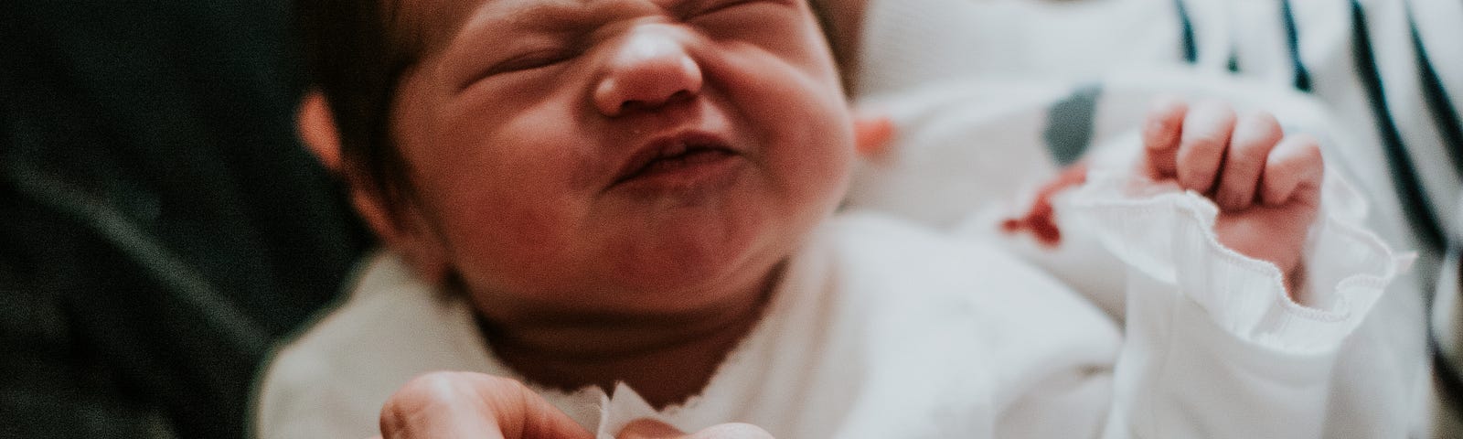 Newborn baby scrunching face at parent who is holding baby’s right hand.