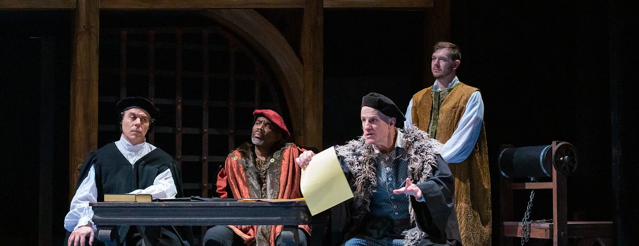 Three men in 16th century robes and hates in black and red sit at a table while the man on the right holds out a piece of parchment paper. A man in a gold robe stands behind them.