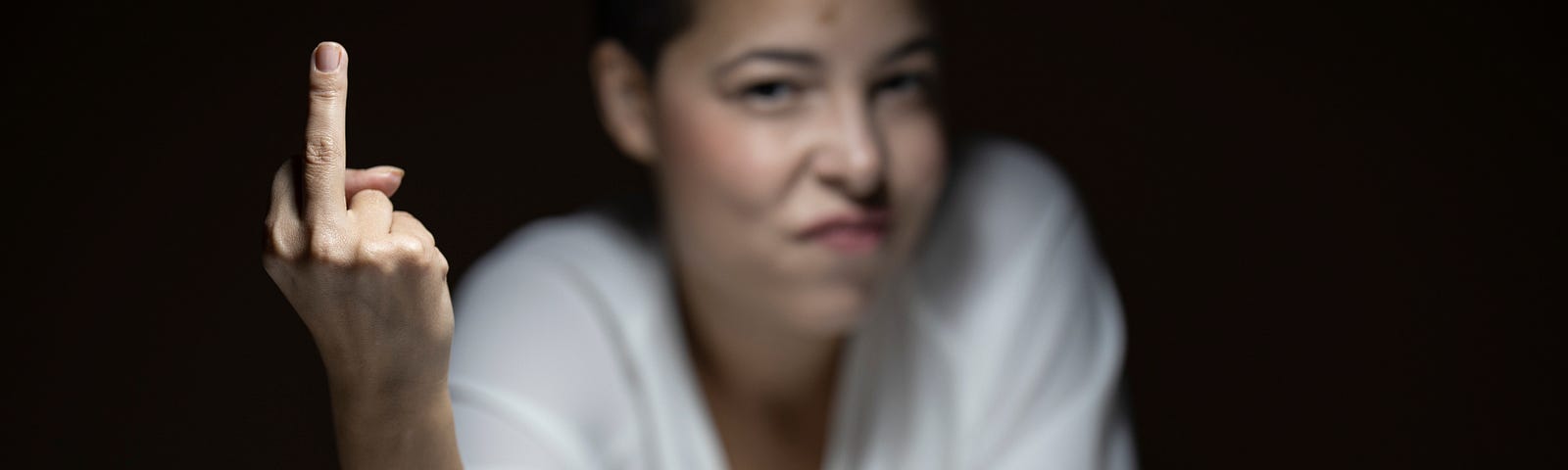 (ID: A white woman is sitting at a table, wearing a white shirt. She is out of focus, except for her right hand, which is giving the middle finger in an angry gesture.)