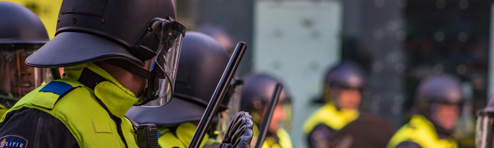 A row of riot cops with full gear, shields, batons, helmets, all dressed in black with some lime green accent on their uniforms, as if ready to fight.