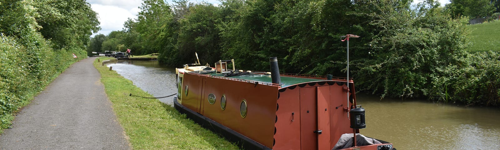 A moored narrowboat on a canal with a broad towpath.