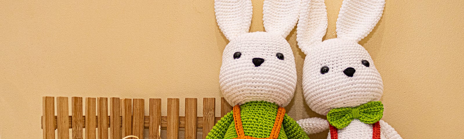 Two crocheted white bunnies in overalls against pastel background