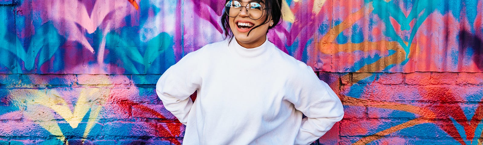 A girl with glasses smiling in front of the colorful graffiti wall