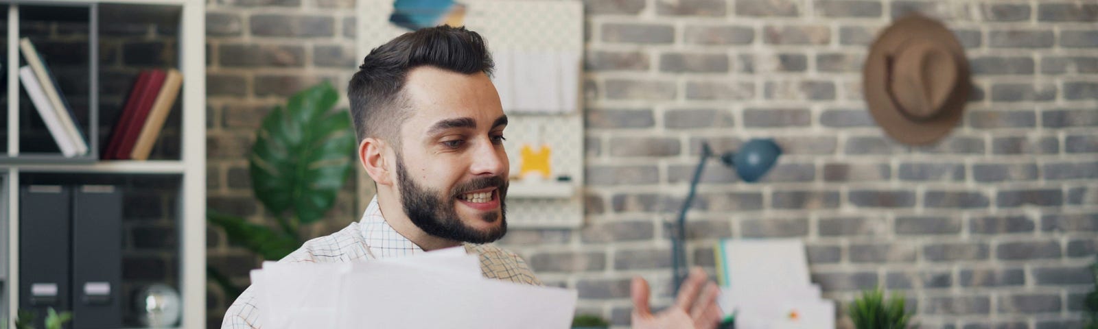 A frustrated man with a beard is sitting at a desk, holding papers and gesturing animatedly in a modern home office.