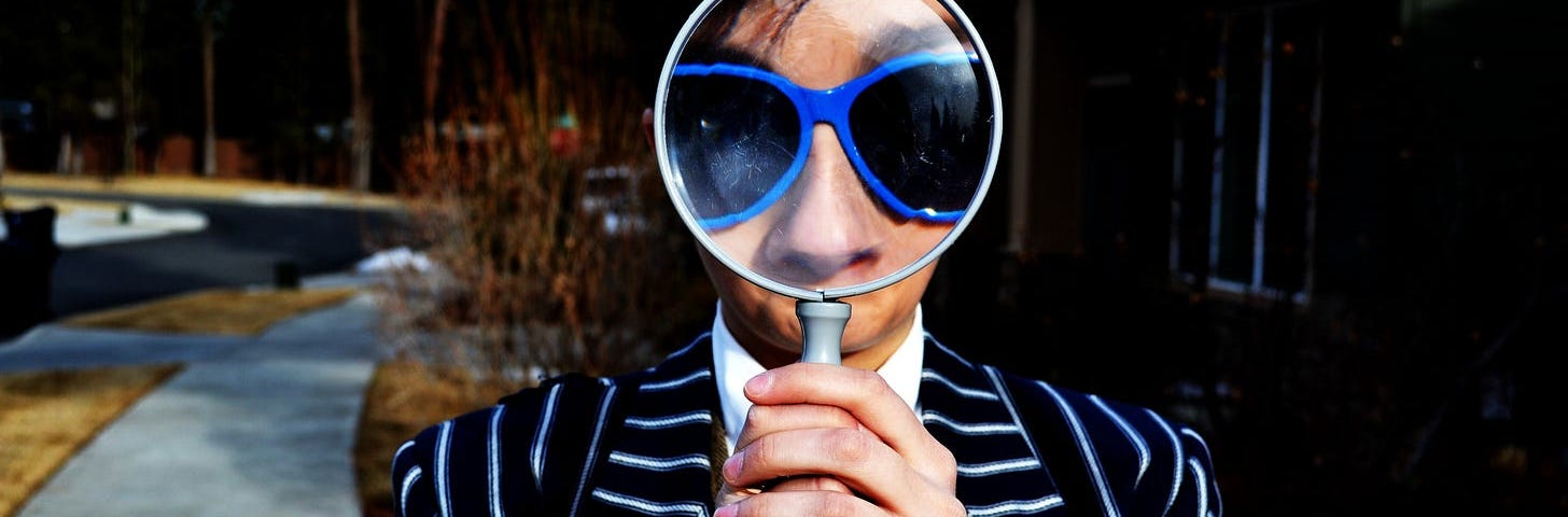 A man in a blue-striped black suite holding up a magnifying glass. He is wearing a blue-rimmed pair of sunglasses.