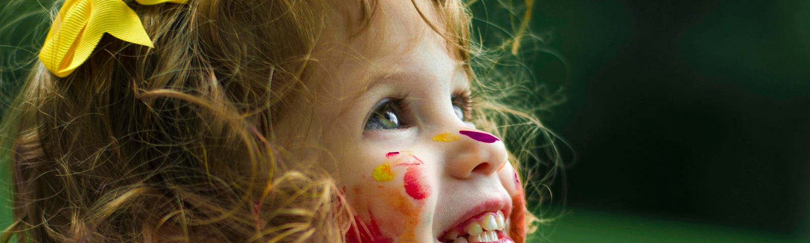 A young child with curly hair and a yellow bow, smiling joyfully with colorful paint smeared on her face and clothes, symbolizing childlike excitement and creativity.