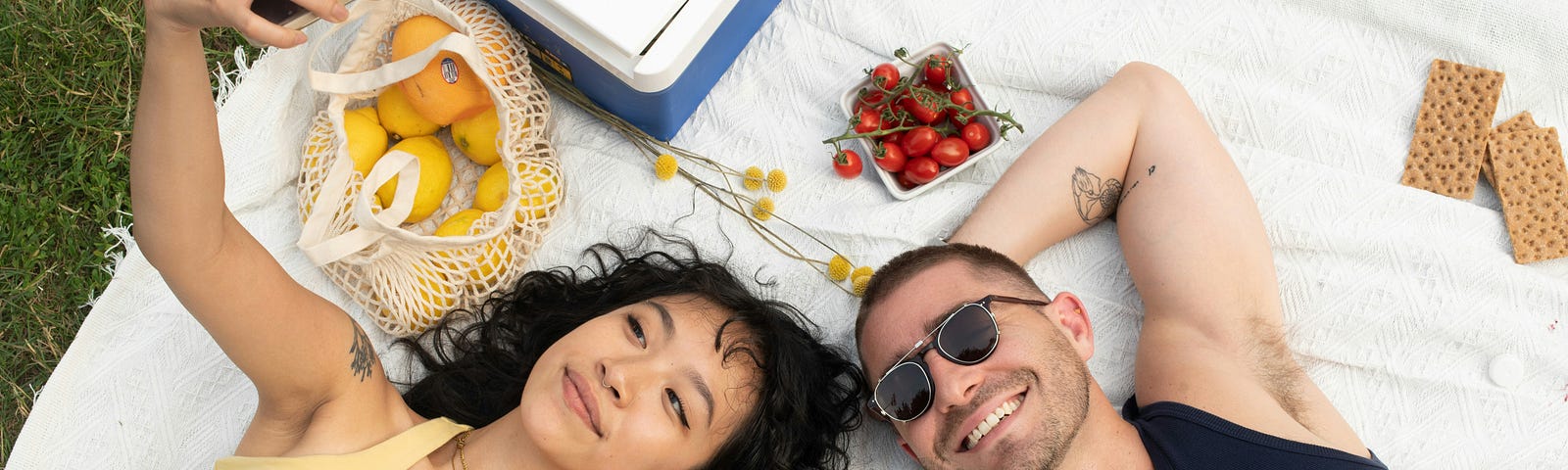 A young couple seem to be njoying taking a selfie laying on a p;icnic blanket with food.