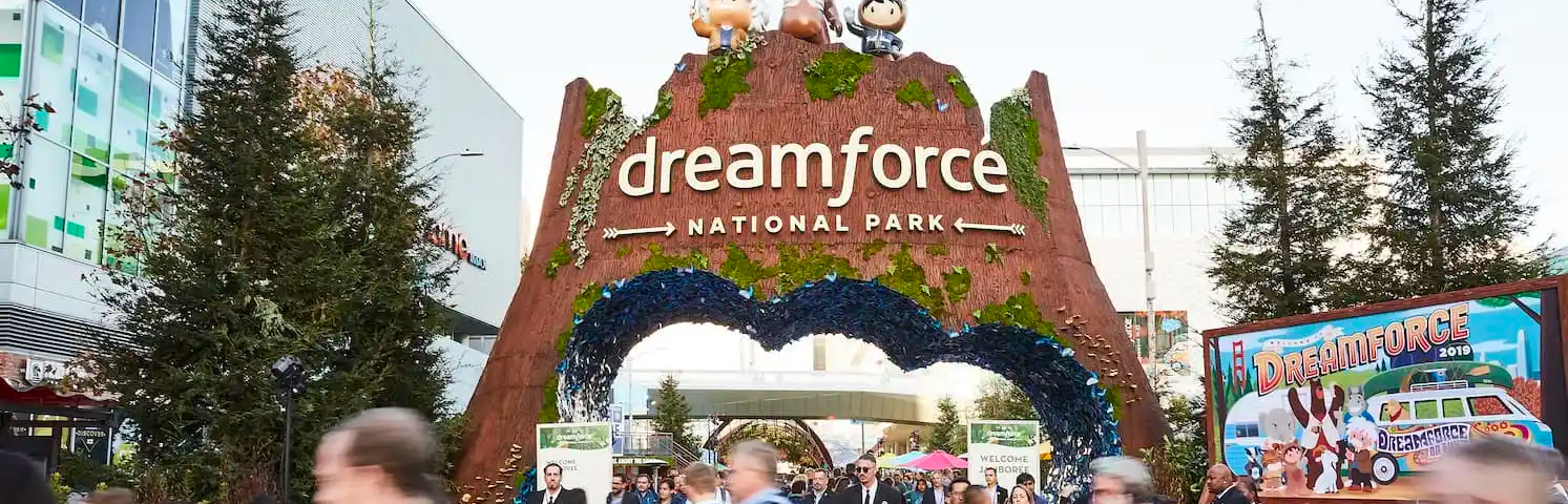 A large structure shaped like a canyon wall that represents the Dreamforce National Park entrance. There’s a cloud-shaped arch cutout. There are lots of people walking through the entrance and also crossing the street in the foreground.