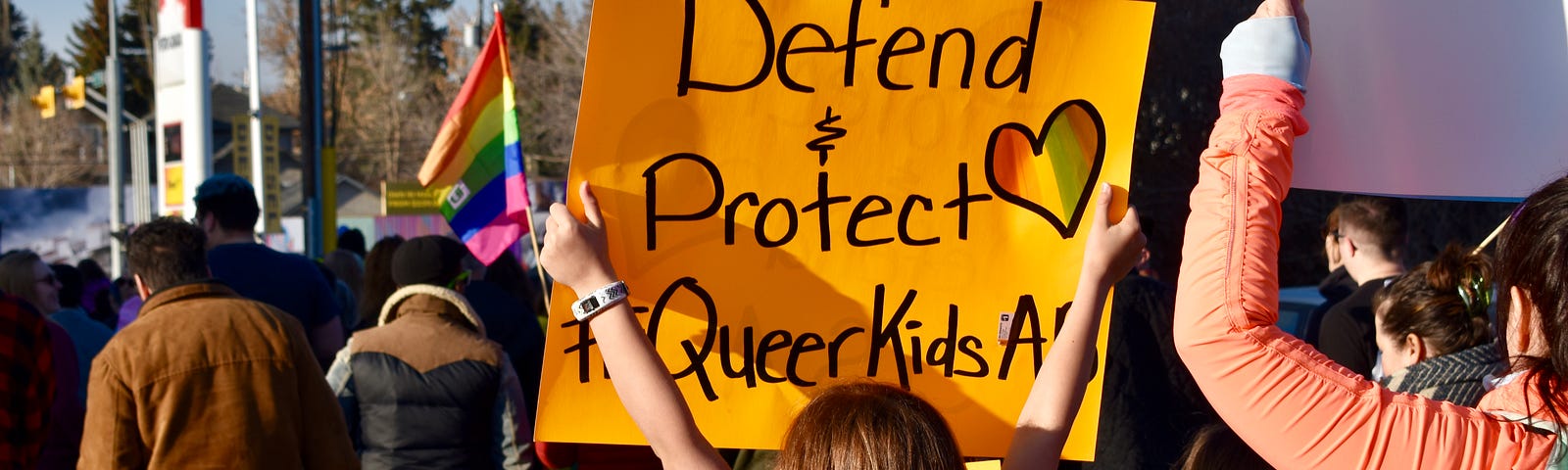 A protest. A child lifts a sign reading, “Defend & Protect Queer Kids”