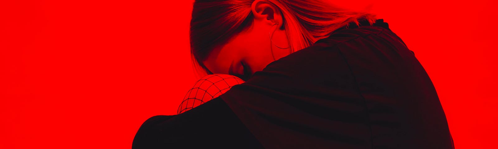 A depressed-appearing young woman sits, her head down while she embraces her bent lefts. Red background.