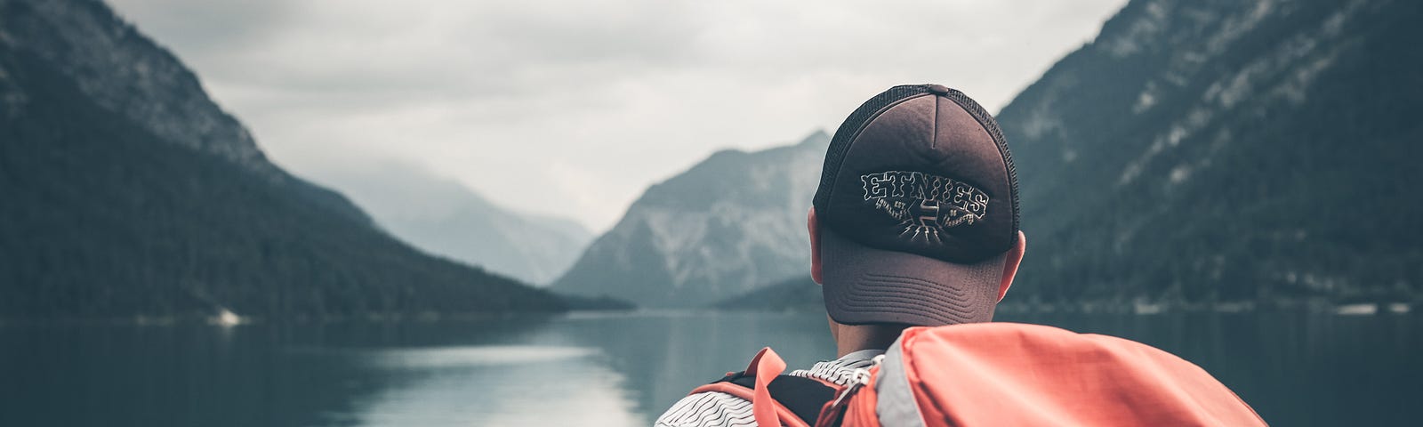 A man with a hiking backpack stares at a lake surrounded by mountains