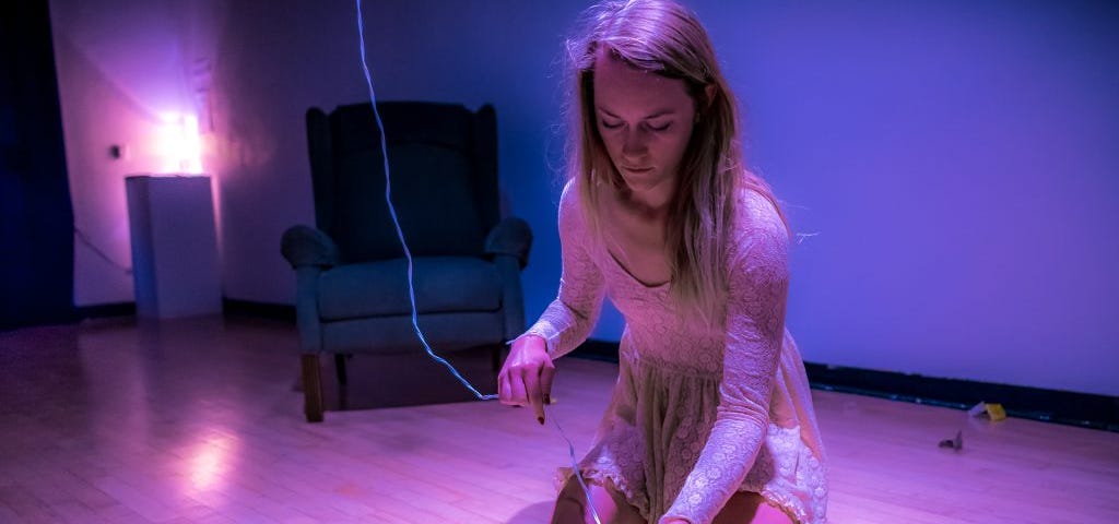 Performance Artwork by Cortnee Brush. Brush is wearing a white, lace dress, and is kneeling on the gallery floor. She holds a vibrator in her hand. The vibrator has a cord that goes up to the gallery ceiling.