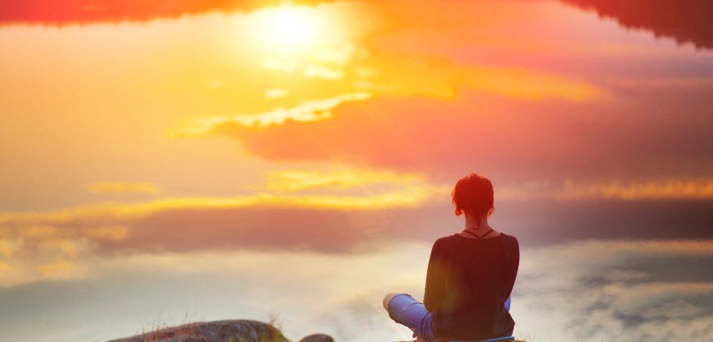 Photo of a woman sitting on a hill above the clouds looking at the sun.