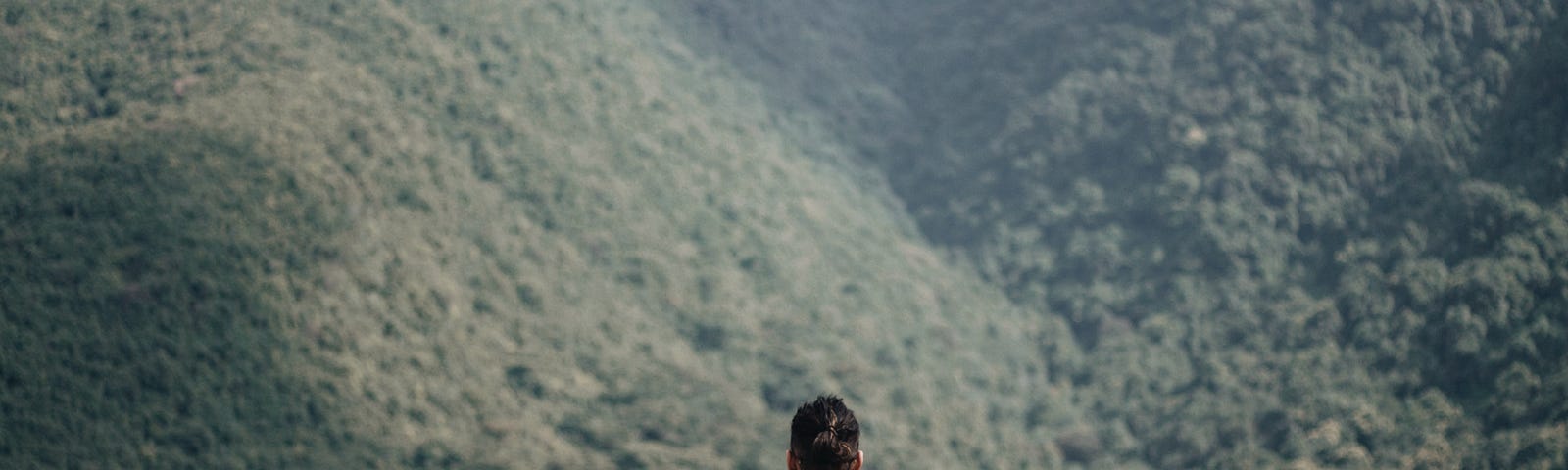 A man with black hair in a bun wearing a checkered blue and black tee sitting on a cliff ledge in front of a forested mountain range. The man has a black backpack sitting on his left side and his phone on his right side.