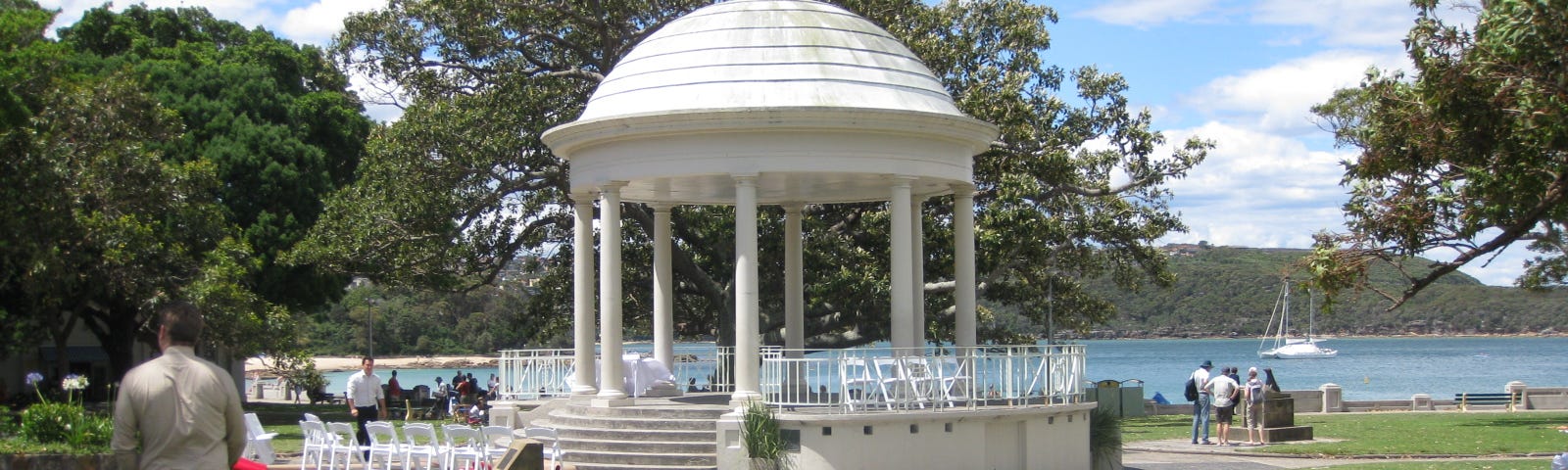 A large, raised rotunda with a white, dome shaped roof, in a grassy waterside park with a boat moored in the distance.