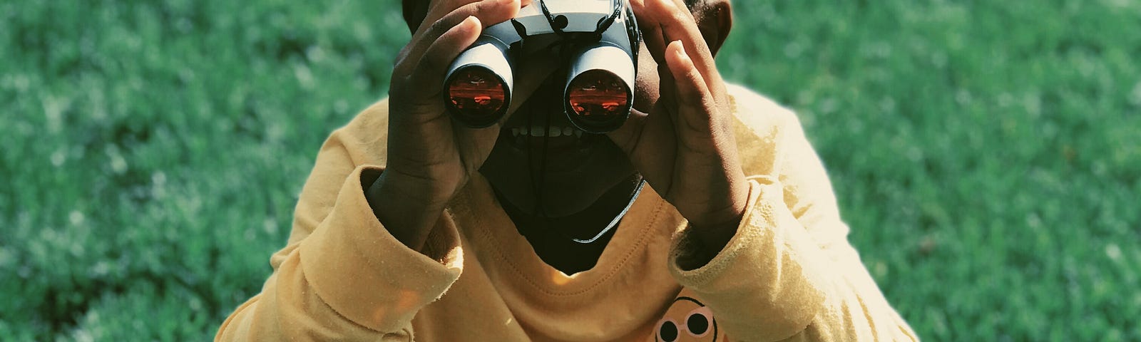 an image of a little black girl sitting cross-legged on a grassed area looking through a tiny pair of binoculars with a broad grin on her face. She is wearing black and white striped tights and a yellow long sleeve shirt. She appears happy and excited at whatever she is viewing.