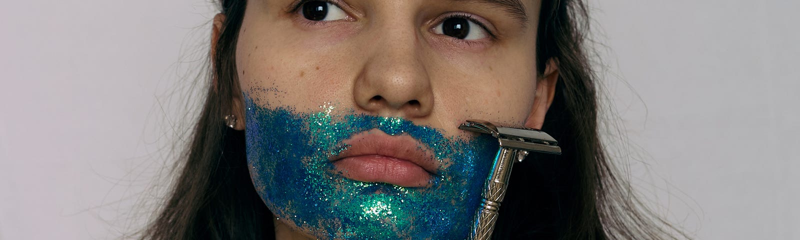 A woman with a beard and mustache made out of blue glitter holds a shaving razoe next to her face in a thoughtful moment.