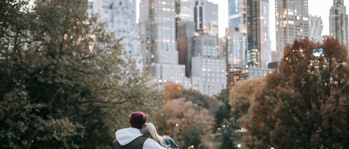 A couple is sitting on a large rock looking at a skyline with trees in front.