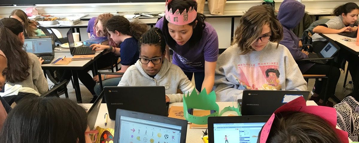 Classroom of students working on Scratch using personal computers. A teacher in pink crown assists students in the upper left side of the image.