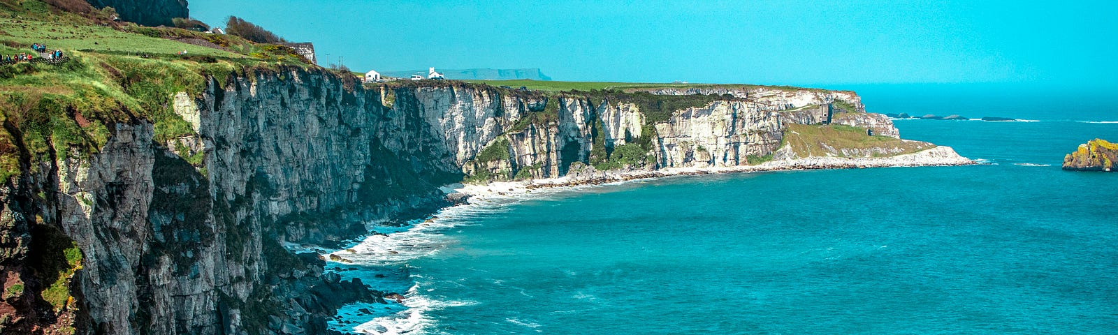 This is a picture of the Irish sea cliffs on a bright, clear day. The top of the cliffs is a vibrant green, and there is white seafoam near the rocks below. I am unsure if these are part of the Cliffs of Moher or not.