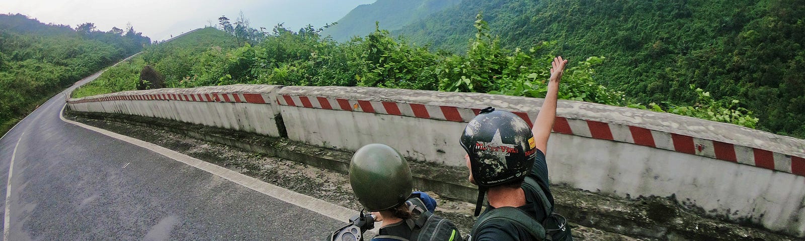 Two people riding a scooter away from the camera on a mountain road. The photograph appears to have been taken by the pillion passenger who can be seen holding a selfie stick.