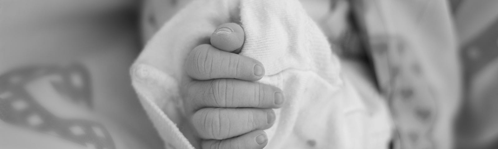 Black and white photo with closeup of an infant’s hand in an oversized sleeve. The baby’s sleeve with hand is surrounded by a blanket with non discernable pattern in gray tones.
