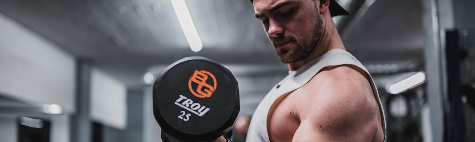 A muscular man in a tank top and baseball cap curls a dumbbell.