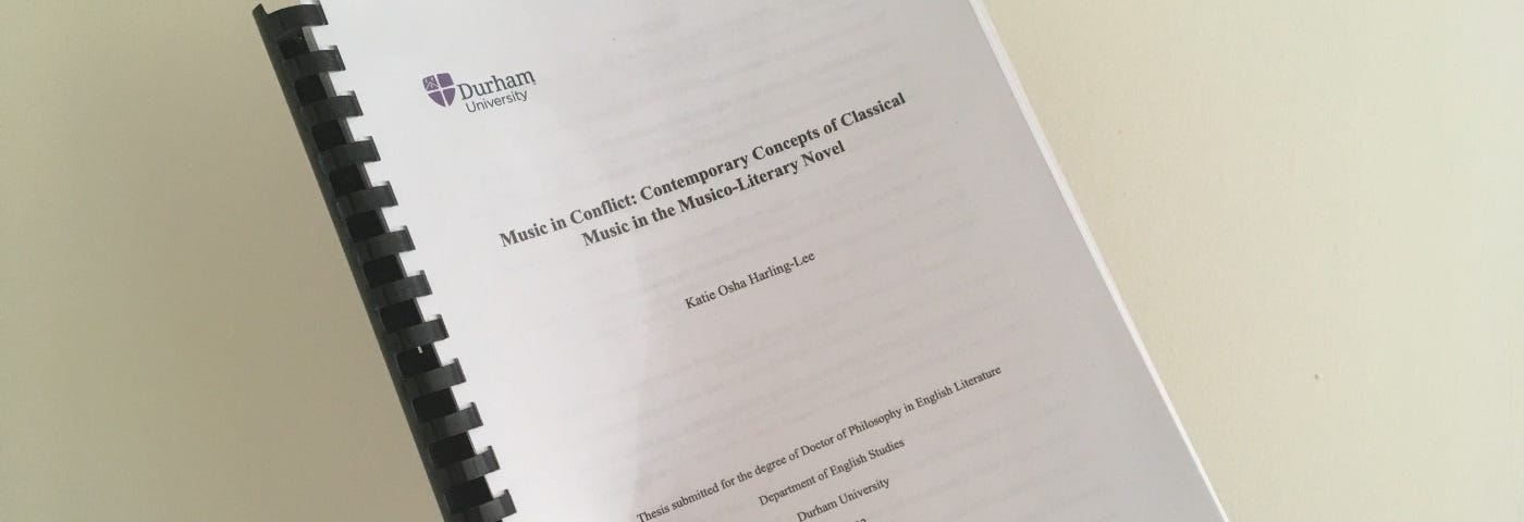Photograph of a ringbound PhD thesis, black text on white paper, the title page visible, held in the air with one white, small hand against a beige wall.