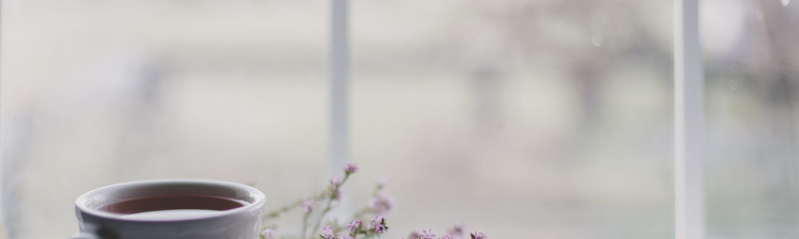 A teacup (and saucer) sit to the right of the screen, the background blurry. Small purple flowers are in the middle of the image. Black tea has flavonols, subtances that can reduce abdominal aortic calcifications.