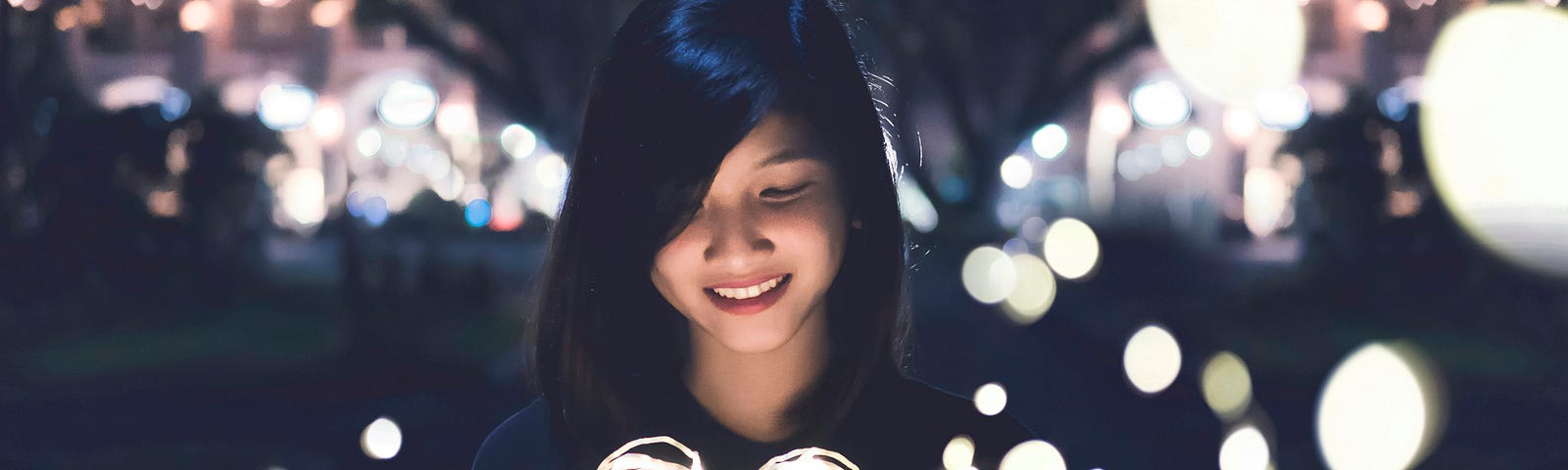 A young woman with a smile on her face holding a lit heart, surrounded by small lights