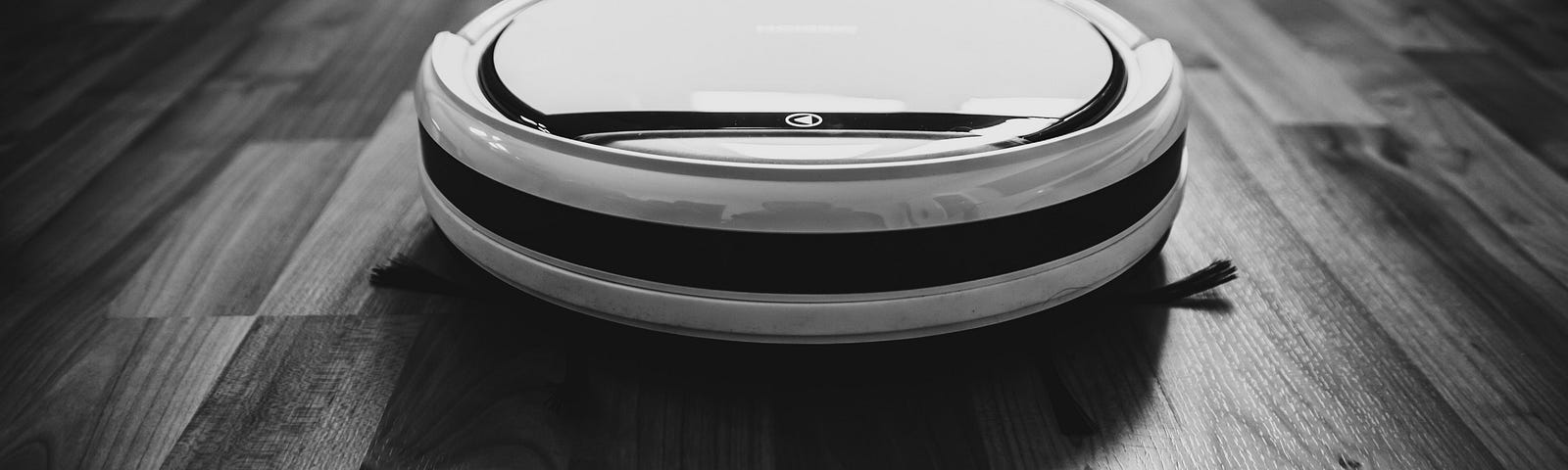 A black and white photo of a robot vacuum cleaner on a wooden laminate floor.