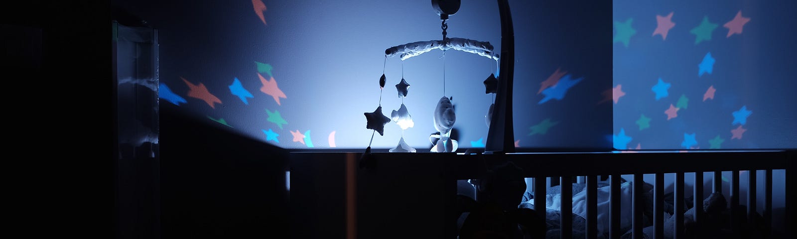 Photo: A crib is visible only as a dark silhouette in a deep blue room. A nightlight projects stars onto the surrounding walls.